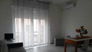 Een moderne woonkamer met een bank, een eettafel met stoelen, een televisie en een raam met lichte gordijnen.