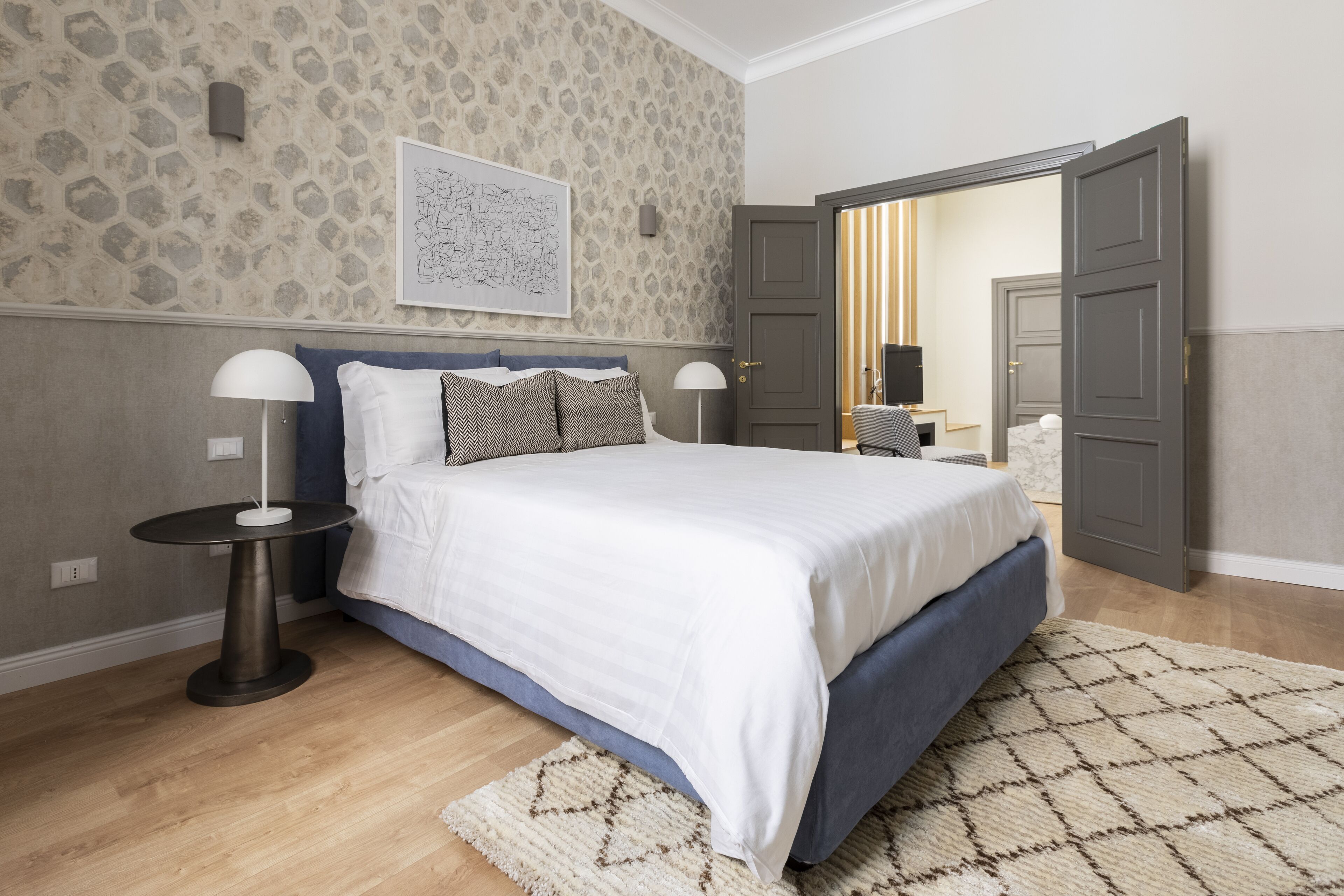 A bedroom with a bed, two bedside tables, a lamp, a rug, and a door leading to another room.