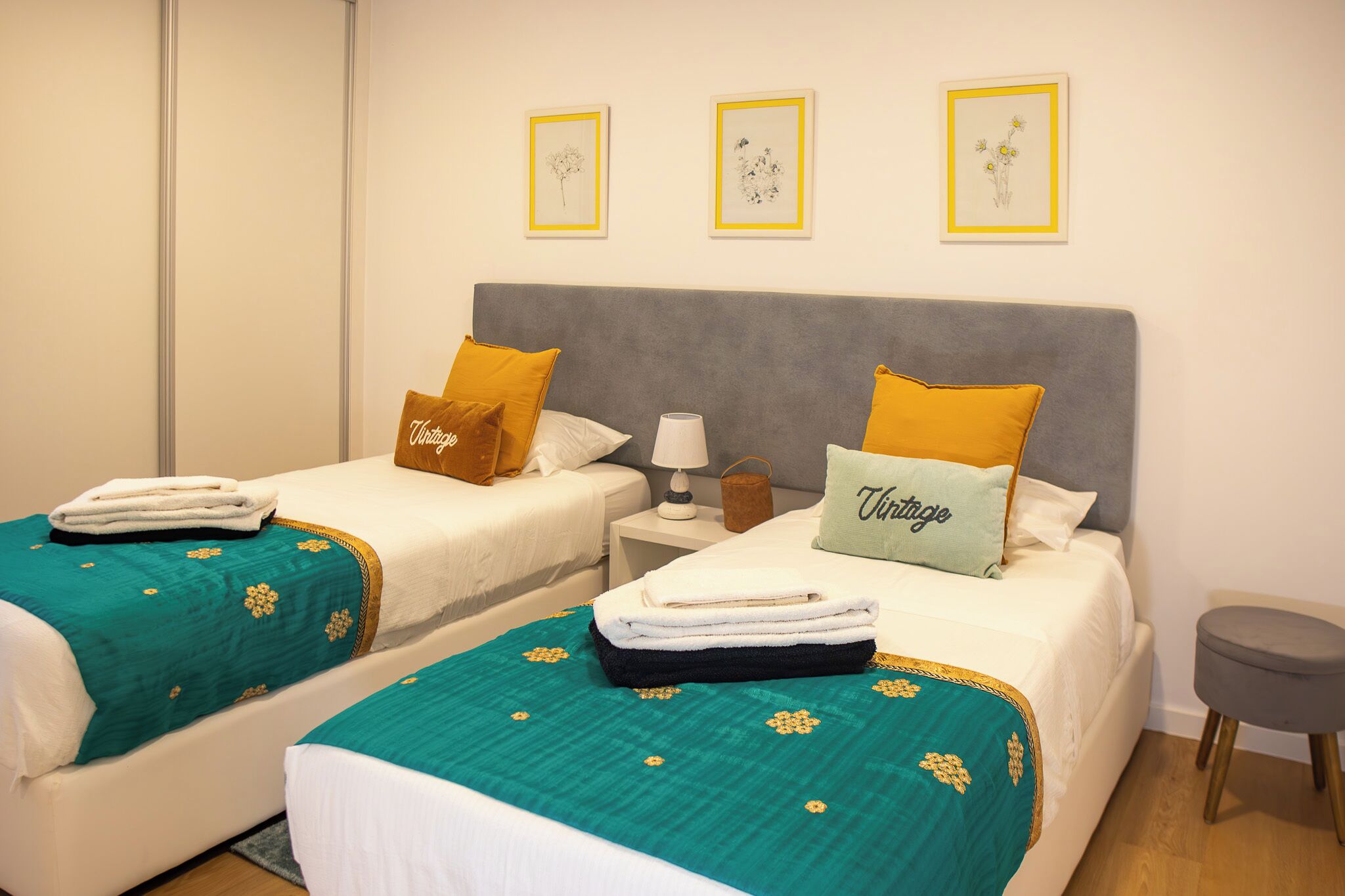 A hotel room with two beds, each with a teal bedspread and white linens, a grey headboard, and a small bedside table with a lamp and a basket.