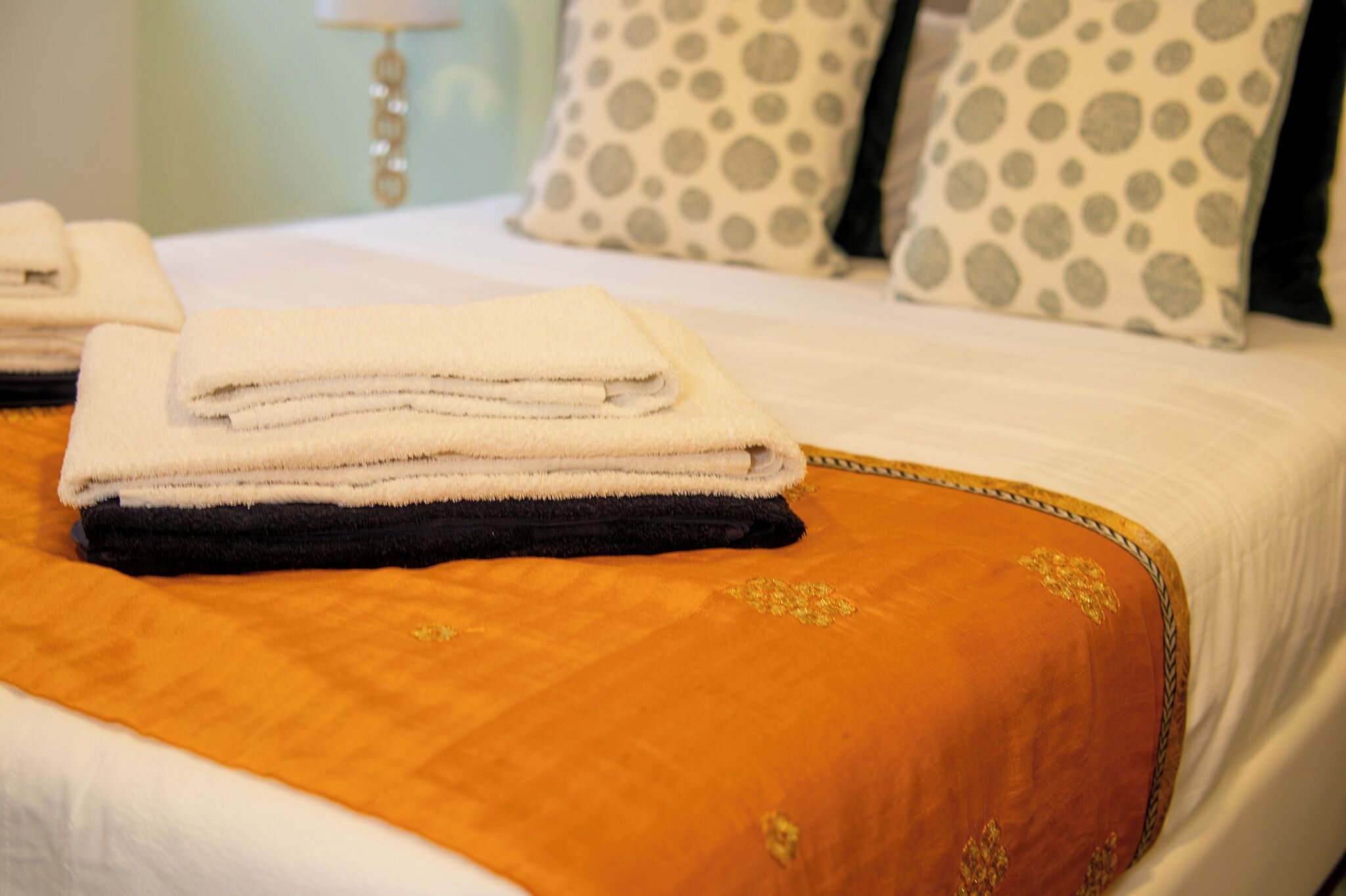 A neatly made bed with a folded white and black towel, a polka dot pillow, and a gold embroidered pillow.