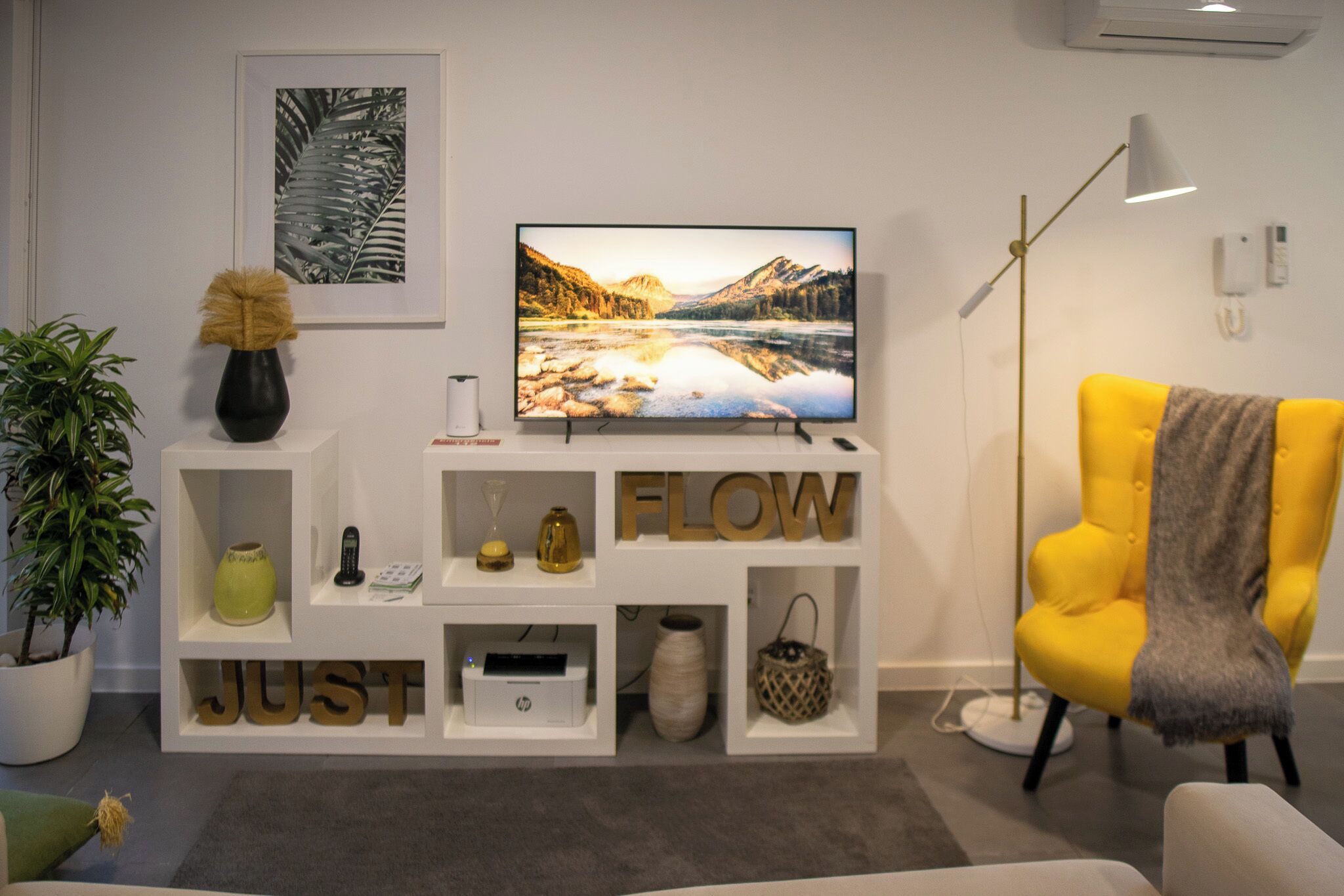 A modern living room with a flat-screen TV, a yellow armchair, and a white shelving unit with decorative items.