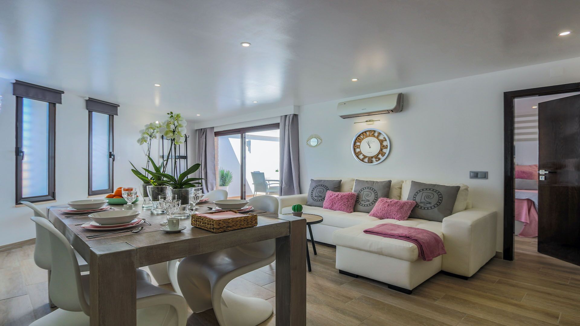 A modern living room with a dining area, featuring a sofa set, a dining table with chairs, a potted plant, and a wall clock.