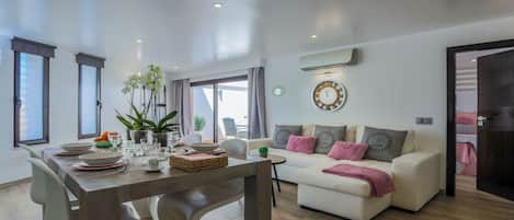 Uma sala de estar moderna com área de jantar, apresentando um conjunto de sofás, uma mesa de jantar com cadeiras, um vaso de plantas e um relógio de parede.