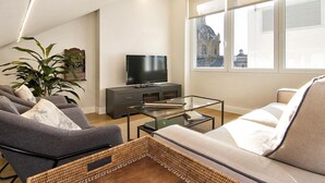 A modern living room with a flat-screen TV, a sofa, a coffee table, and a large window offering a view of a building.