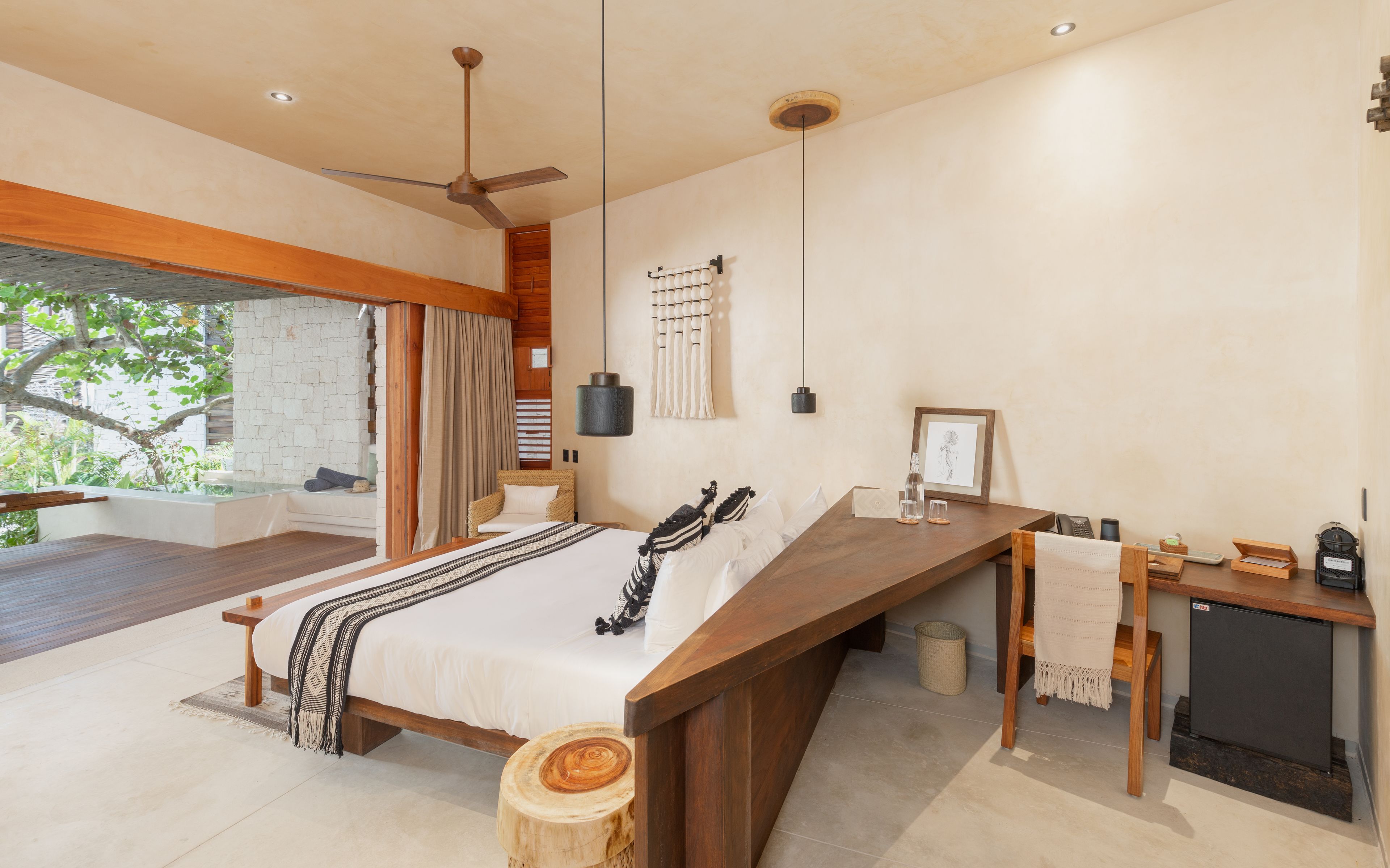 A modern bedroom with a large bed, wooden headboard, a desk with a computer, and a view of greenery outside.