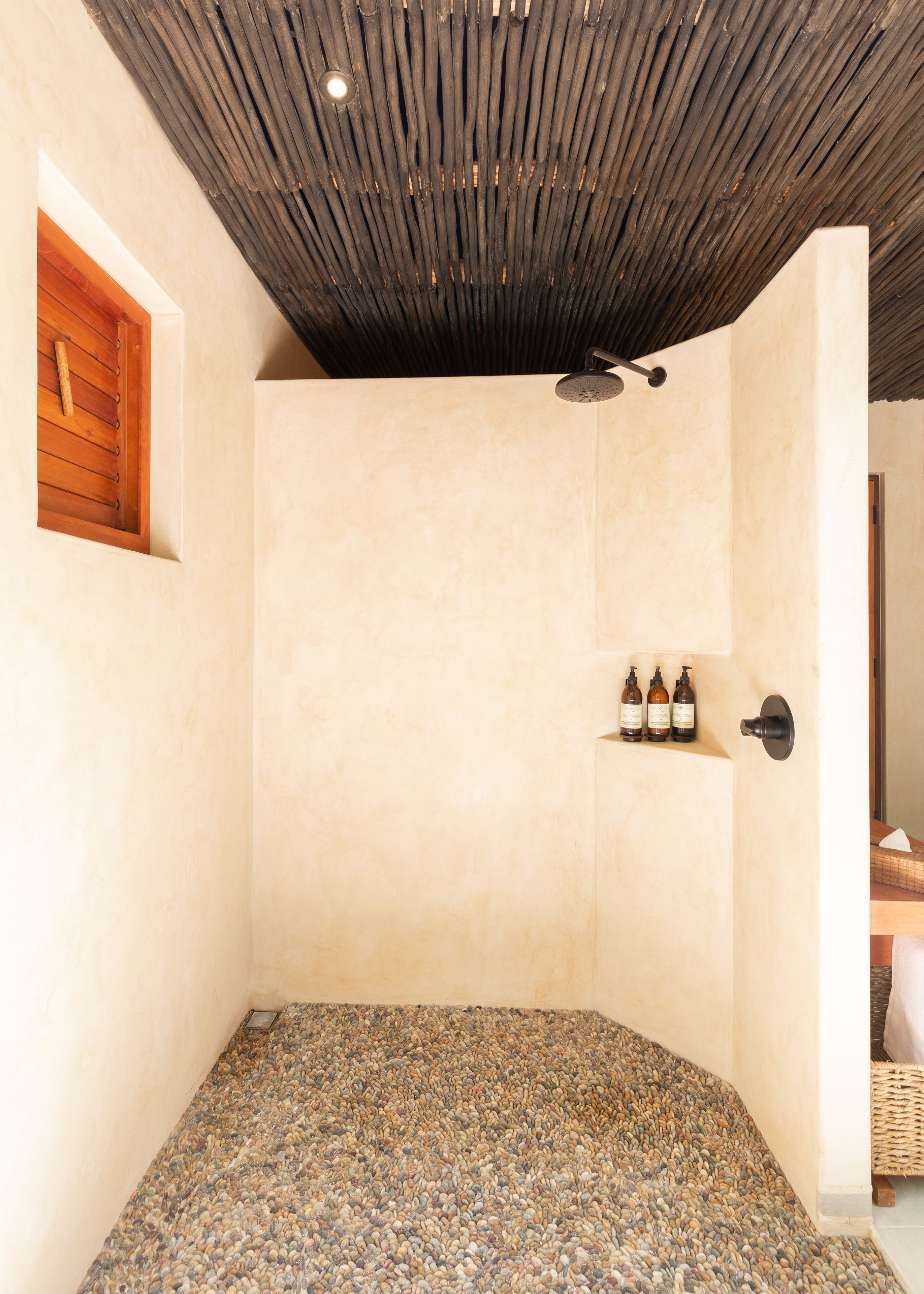 A shower area with a wooden slatted ceiling, a pebble-tiled floor, and a shelf with bottles.