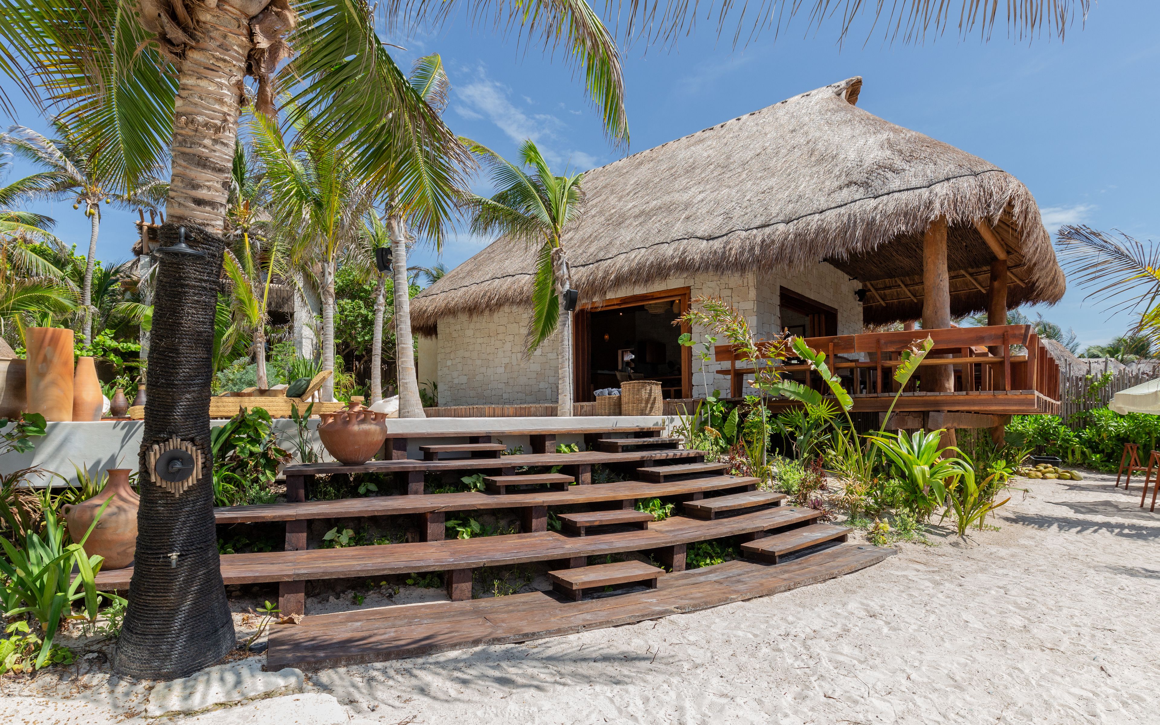 A thatched-roof building with wooden steps leading to a porch, surrounded by tropical vegetation and a sandy beach.