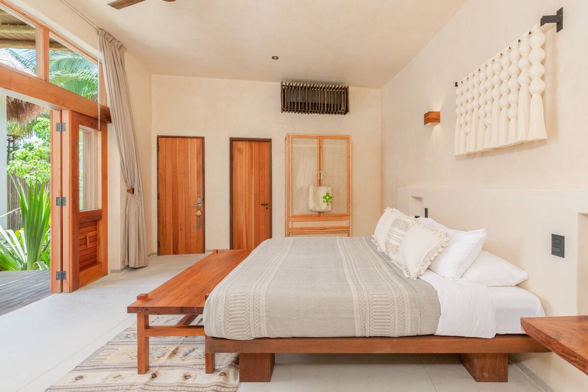 Un dormitorio con una cama de madera, una mesita de noche de madera, un ventanal grande con cortinas y una puerta.
