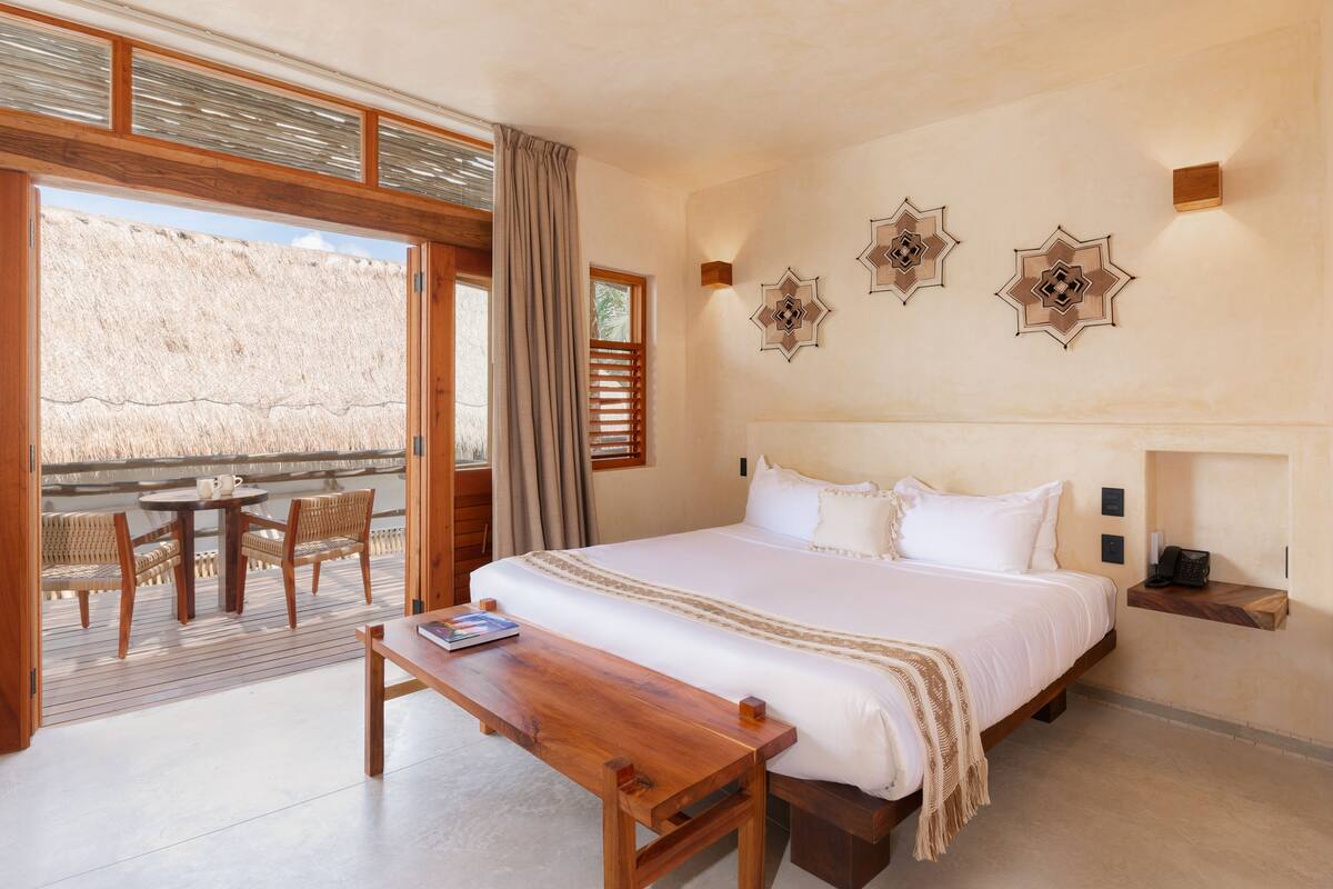 Un dormitorio con una cama grande, un banco de madera, una mesa y un balcón con sillas de mimbre.