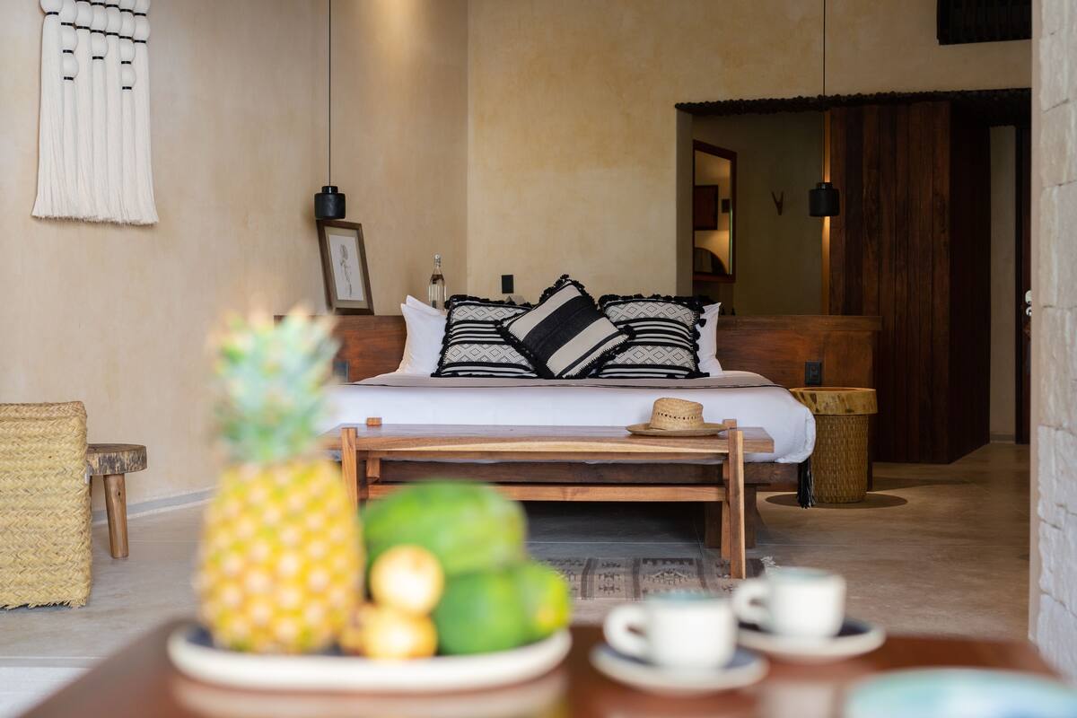 Una habitación de hotel acogedora con cama, mesita de noche y un pequeño rincón de comedor con una piña y un sombrero.