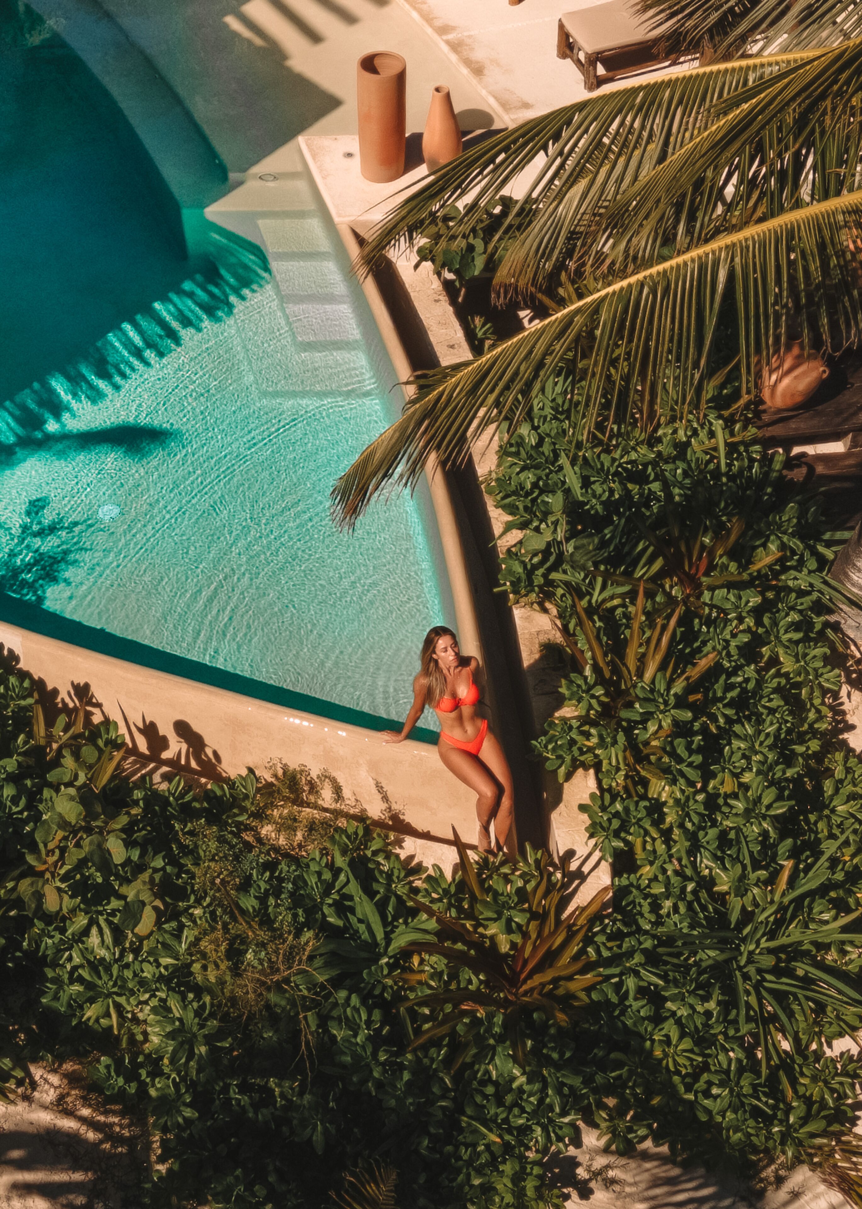 Zona de piscina con una persona en traje de baño, rodeada de exuberante vegetación y una palmera.