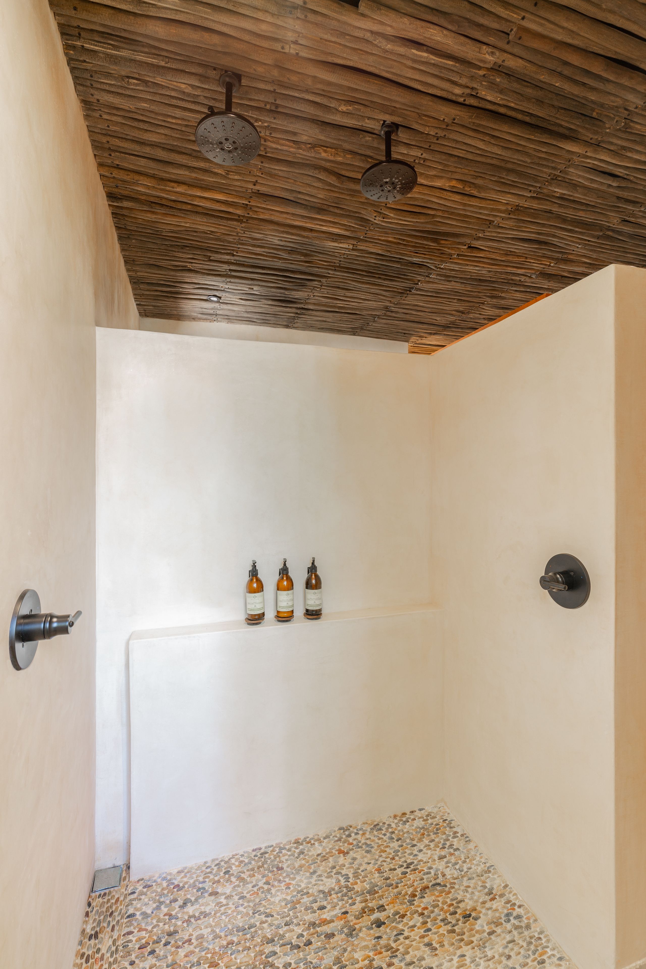 A shower with a wooden ceiling, a white wall, and a tiled floor. There are three bottles on the wall.