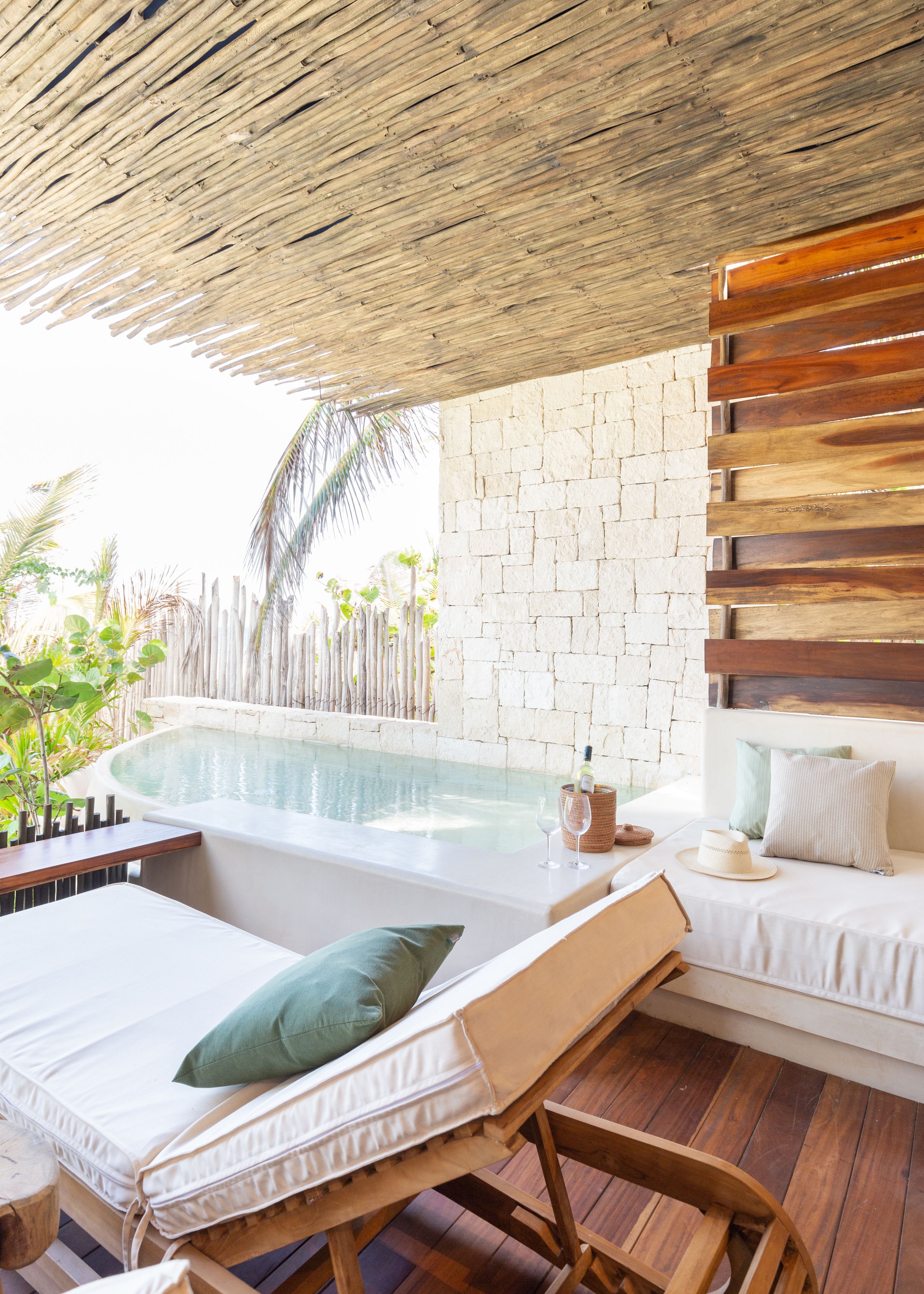 A poolside lounge with a wooden deck, a stone wall, and a cushioned seating area with a bottle of wine and a glass.
