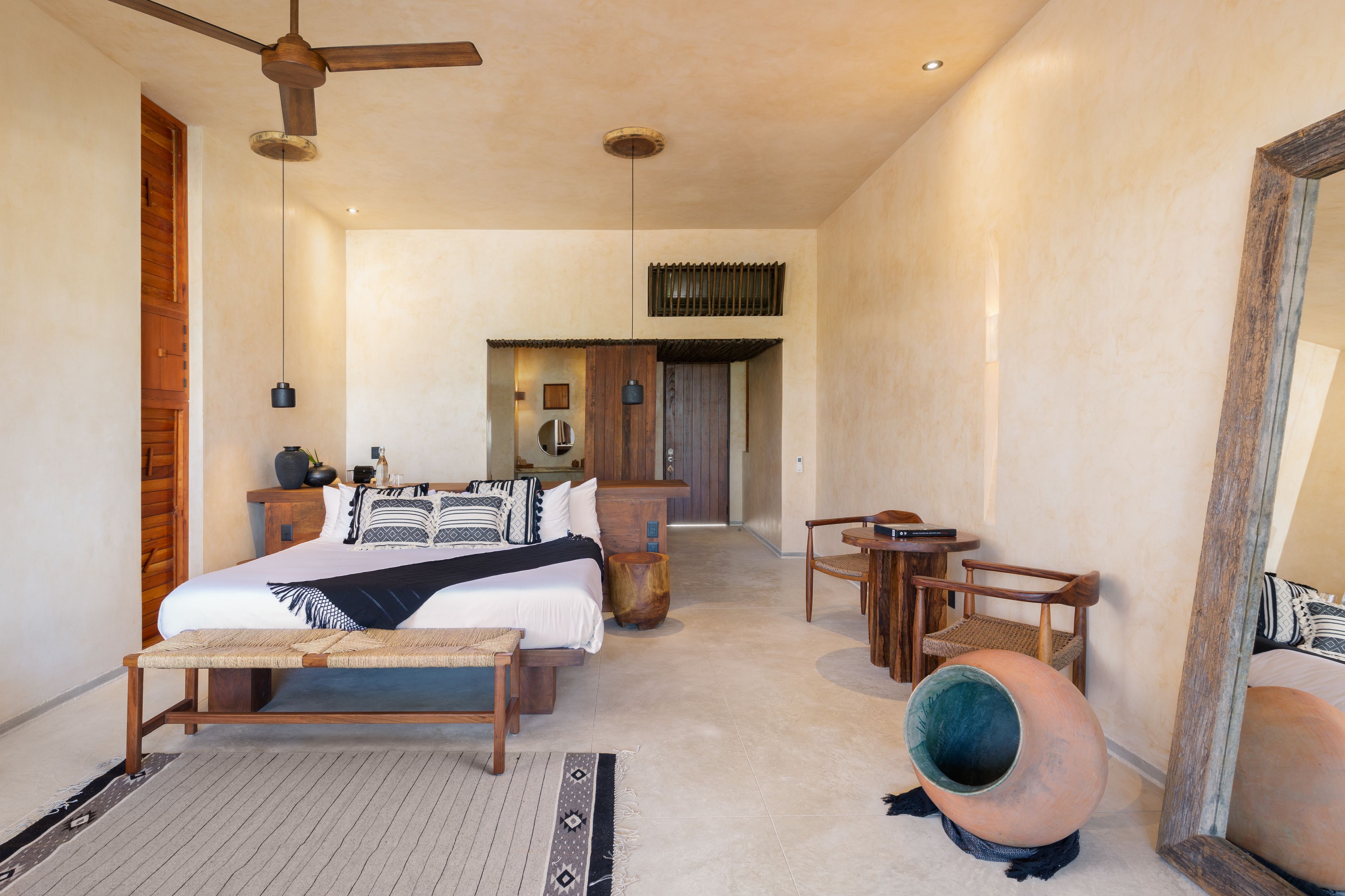 A spacious bedroom with a large bed, a wooden bench, a small table, and a chair. There is a ceiling fan, a mirror, and a large window.