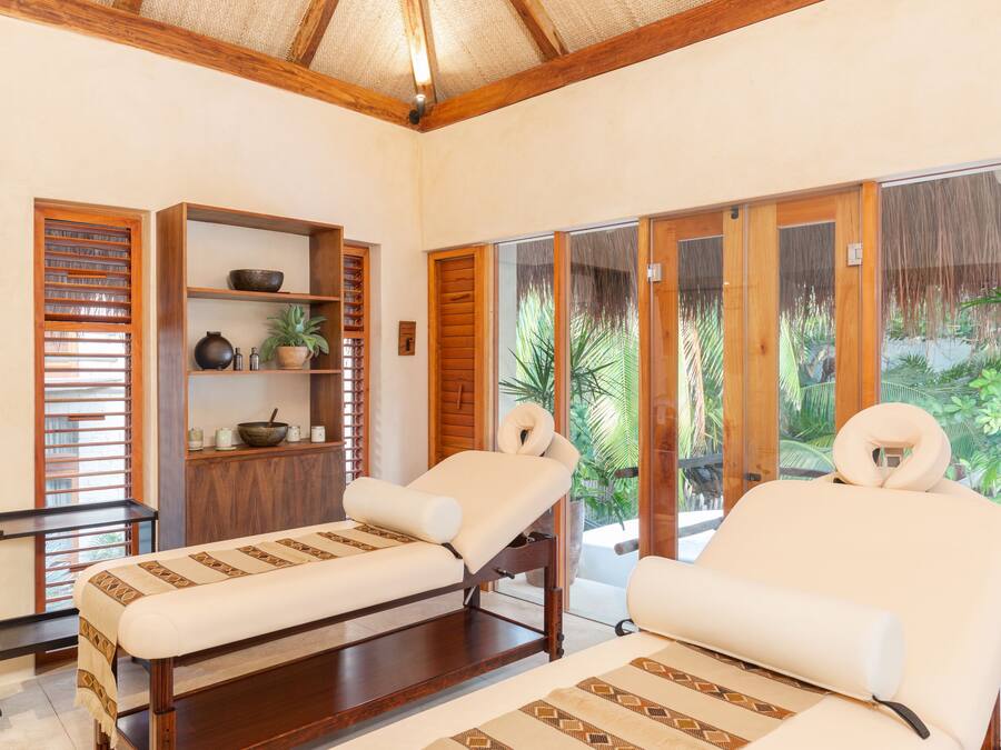 A spa room with two massage tables, wooden shelves, and a large window with a view of greenery.
