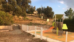 A fenced playground area with a slide and climbing structure, surrounded by trees and a hilly landscape.