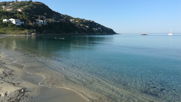 A coastal scene with a clear beach, calm sea, and buildings on a hillside.