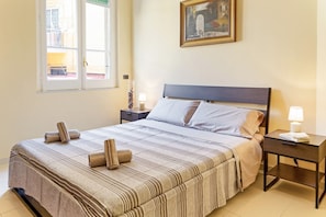 A neatly made bed with striped bedding, a bedside table with a lamp, and a window with a view of buildings.