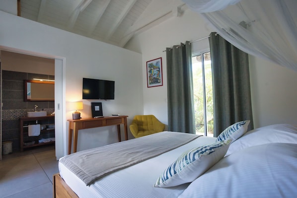 A hotel room with a bed, a desk with a lamp, a chair, a TV, a bathroom with a sink and mirror, and a window with curtains. - Le Cocotel (Saint-François)