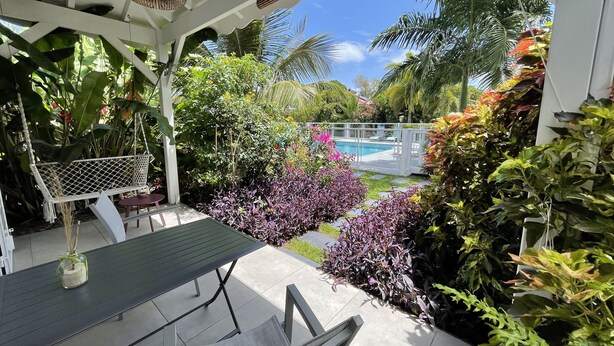 A covered outdoor area with a table and chairs, surrounded by lush tropical plants and a view of a pool. - Le Cocotel (Saint-François)
