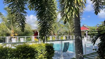 Zona de piscina con barandilla de vidrio, palmeras y un edificio rojo al fondo.