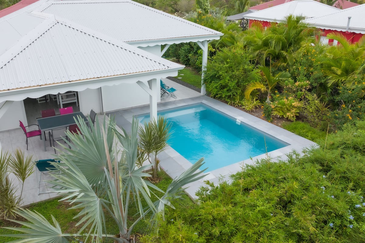 A white house with a pool, surrounded by lush greenery.