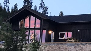Une cabane en bois à deux étages, dotée de grandes fenêtres et d’un toit en pente, entourée d’arbres.