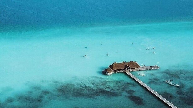 An aerial view of a tropical beach with clear turquoise waters, a thatched-roof hut, and a pier extending into the sea. - Hotel Tuparenda Bacalar (Bacalar)