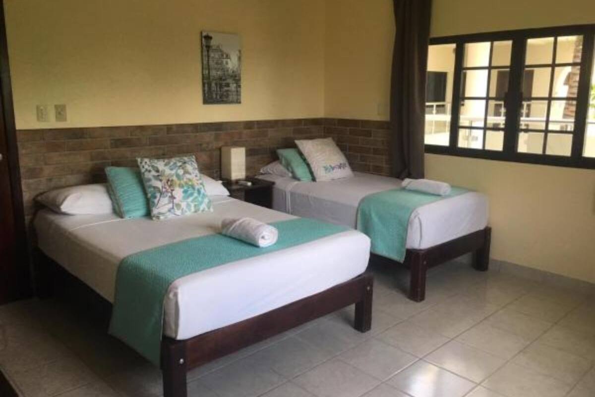 A hotel room with two single beds, each with a green blanket and a pillow. There is a nightstand between the beds, and a window with a view of the outside.