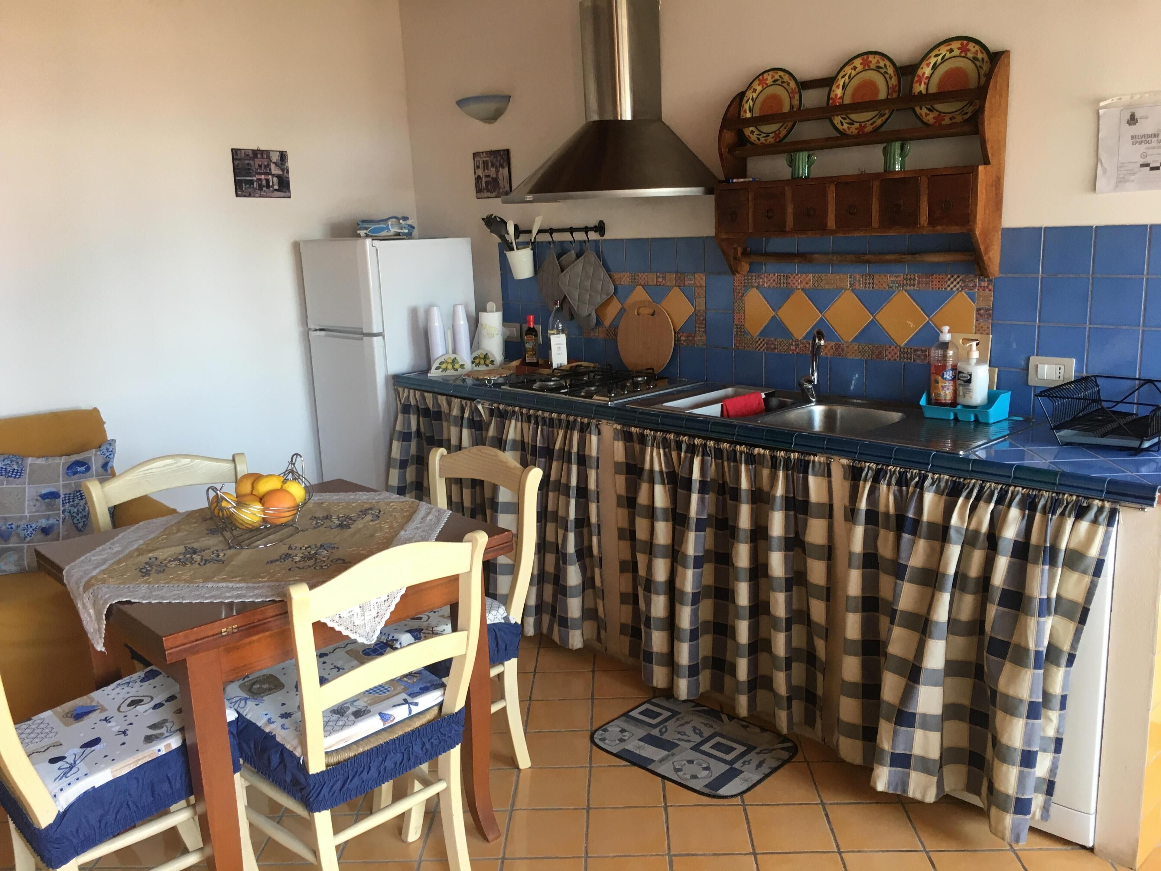 A compact kitchen with a refrigerator, stove, and sink. There are chairs and a table with a fruit bowl.