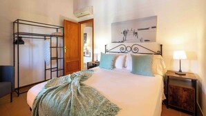 Un dormitorio con cama, mesita de noche, estante en la pared, puerta y un cuadro.