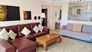 A living room with a red leather sofa, a wooden coffee table, and a light-colored rug. There is a kitchen area with cabinets and a countertop visible in the background. - Villa Essence - with A/C, heated private pool, hottub, gardens & stunning views! (Tias)