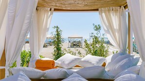 A sunlit room with a bed, white curtains, and a view of a beach and umbrellas.