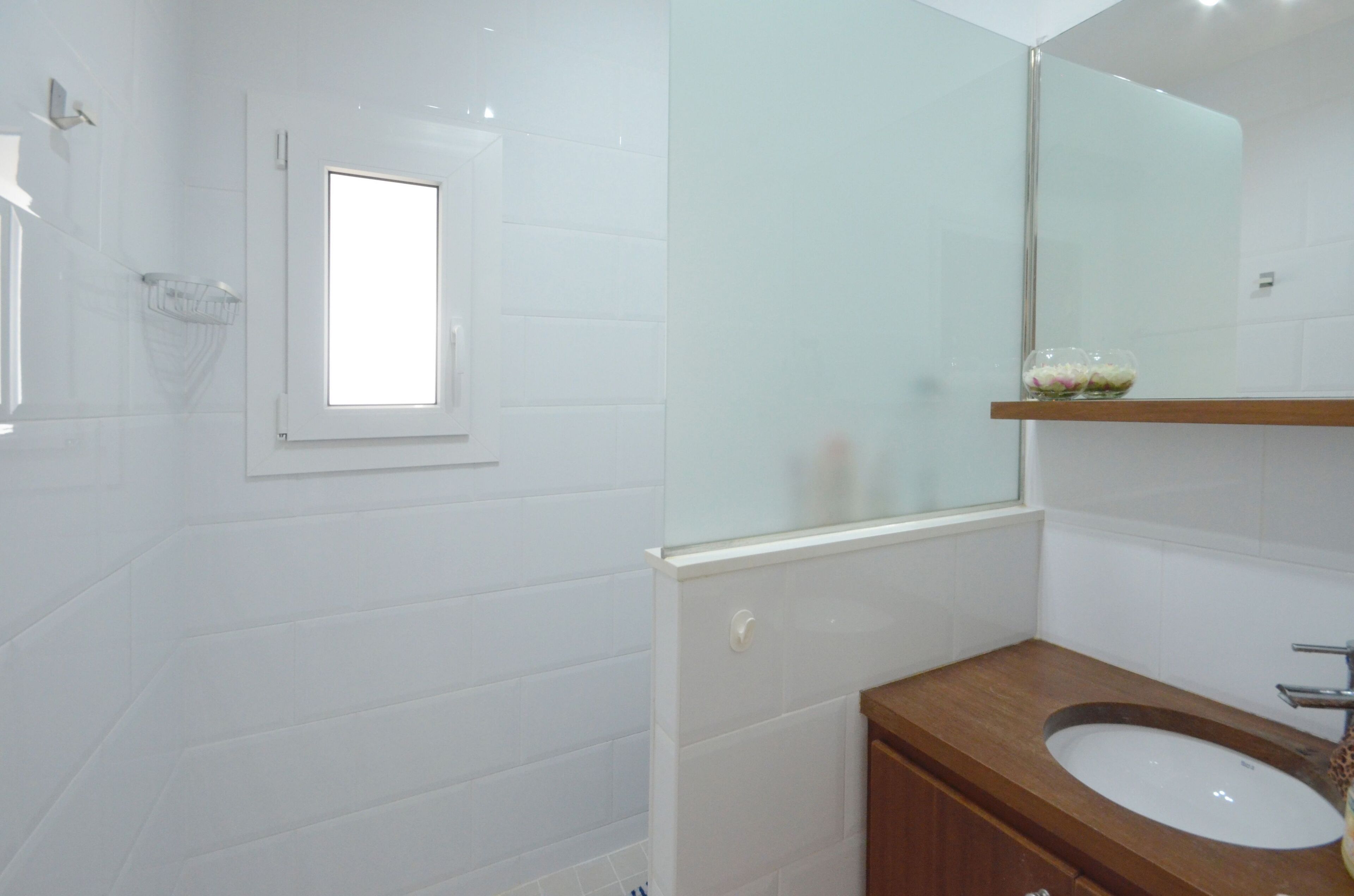 A modern bathroom with white tiles, a wooden vanity, a large mirror, and a small window.