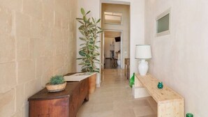 Un pasillo estrecho con un banco de madera a la izquierda, una planta en maceta y una mesa de madera con una lámpara a la derecha.