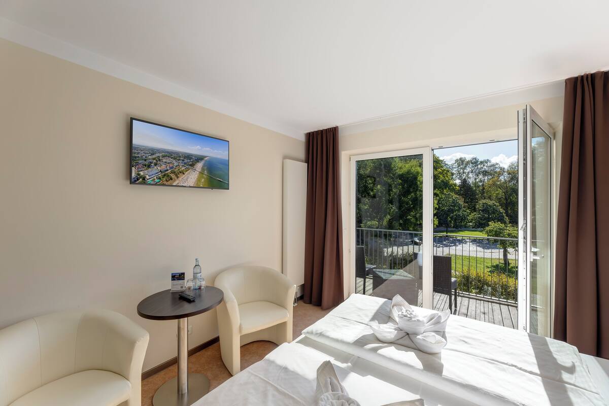 A hotel room with a bed, a chair, a small table, a painting, and a balcony with a view of greenery and a parking area.