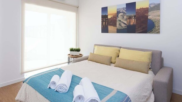 A hotel room with a large bed, a window with blinds, a small table with a plant, and a wall-mounted artwork. - Anna´s Flats by HostWise (Vila Nova de Gaia)