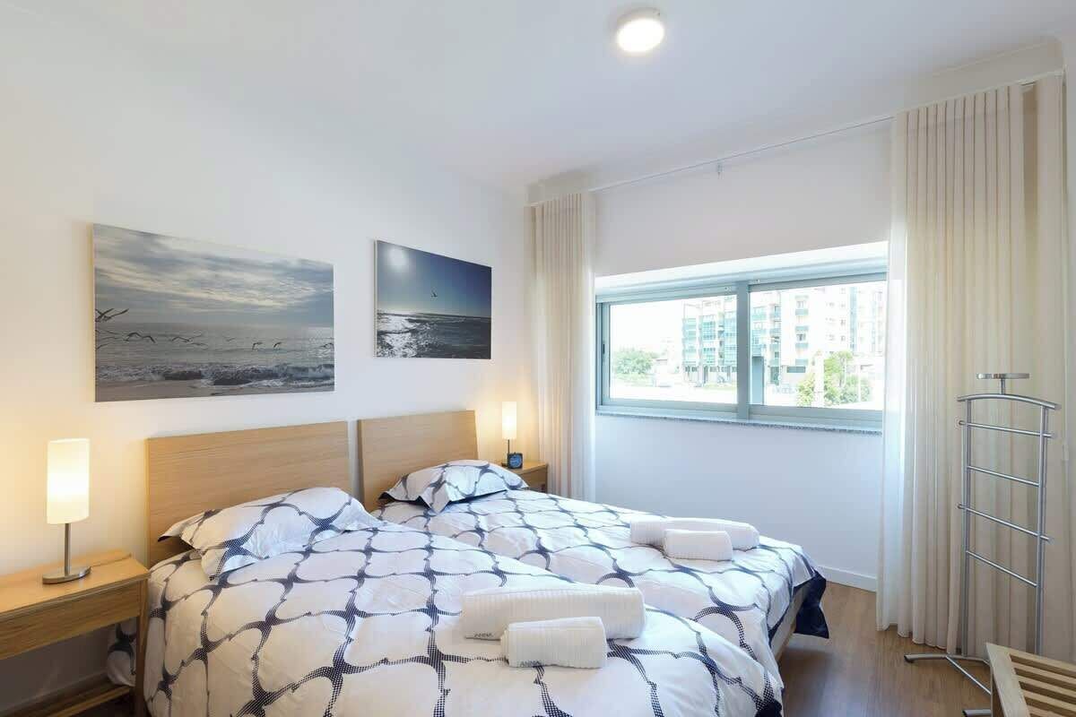 Una habitación de hotel con una cama, dos cuadros colgados en la pared, una ventana con cortinas, una mesita de noche con lámpara y un perchero para toallas.
