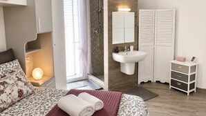 A bedroom with a bed, a bedside lamp, a bathroom with a shower, a sink, and a mirror. - New Construction,  beautiful 2 bed apartment facing the sea, Wi-fi ,Aircon (La Ciaccia)