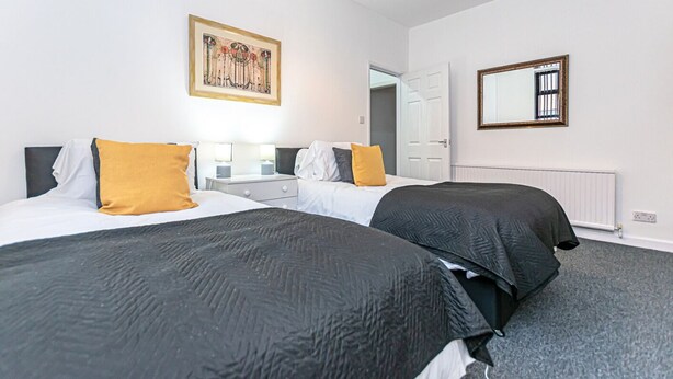 A hotel room with two beds, a nightstand, a mirror, and a painting on the wall. - Spacious 3 Bedroom House in Wigan – Perfect for Contractors & Families (Hindley)