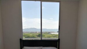 A bedroom with a large window, a bed with white bedding, and a view of greenery and distant buildings. - Exclusive Vacation Apartment in Peñalisa (Ricaurte)