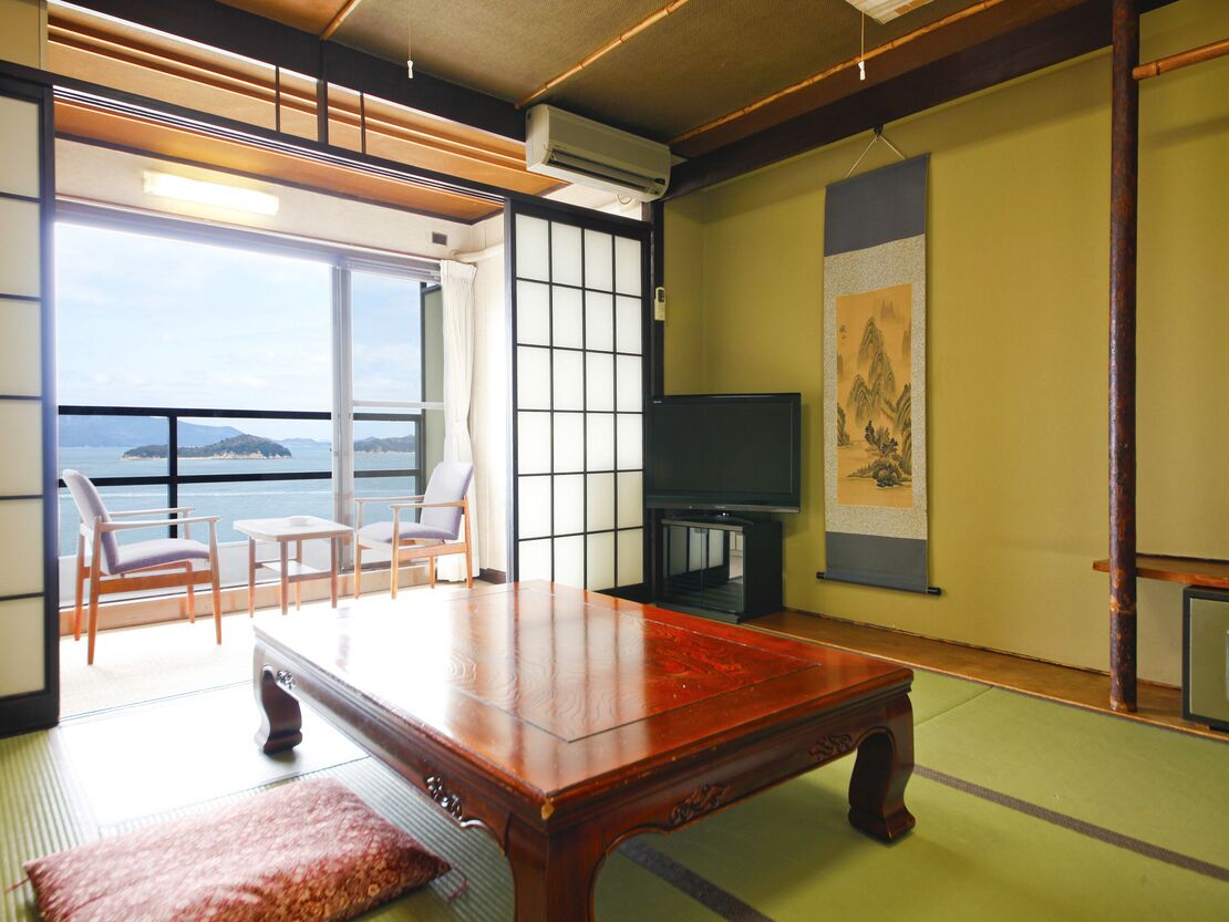 A traditional Japanese room with tatami flooring, a low wooden table, a television, and a view of the sea.