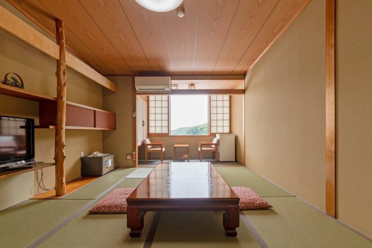 Ein traditioneller japanischer Raum mit Tatami-Boden, einem niedrigen Tisch, einem Fernseher und Blick auf einen Berg.