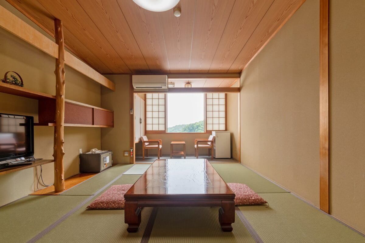 A traditional Japanese room with tatami flooring, a low table, a television, and a view of a mountain.