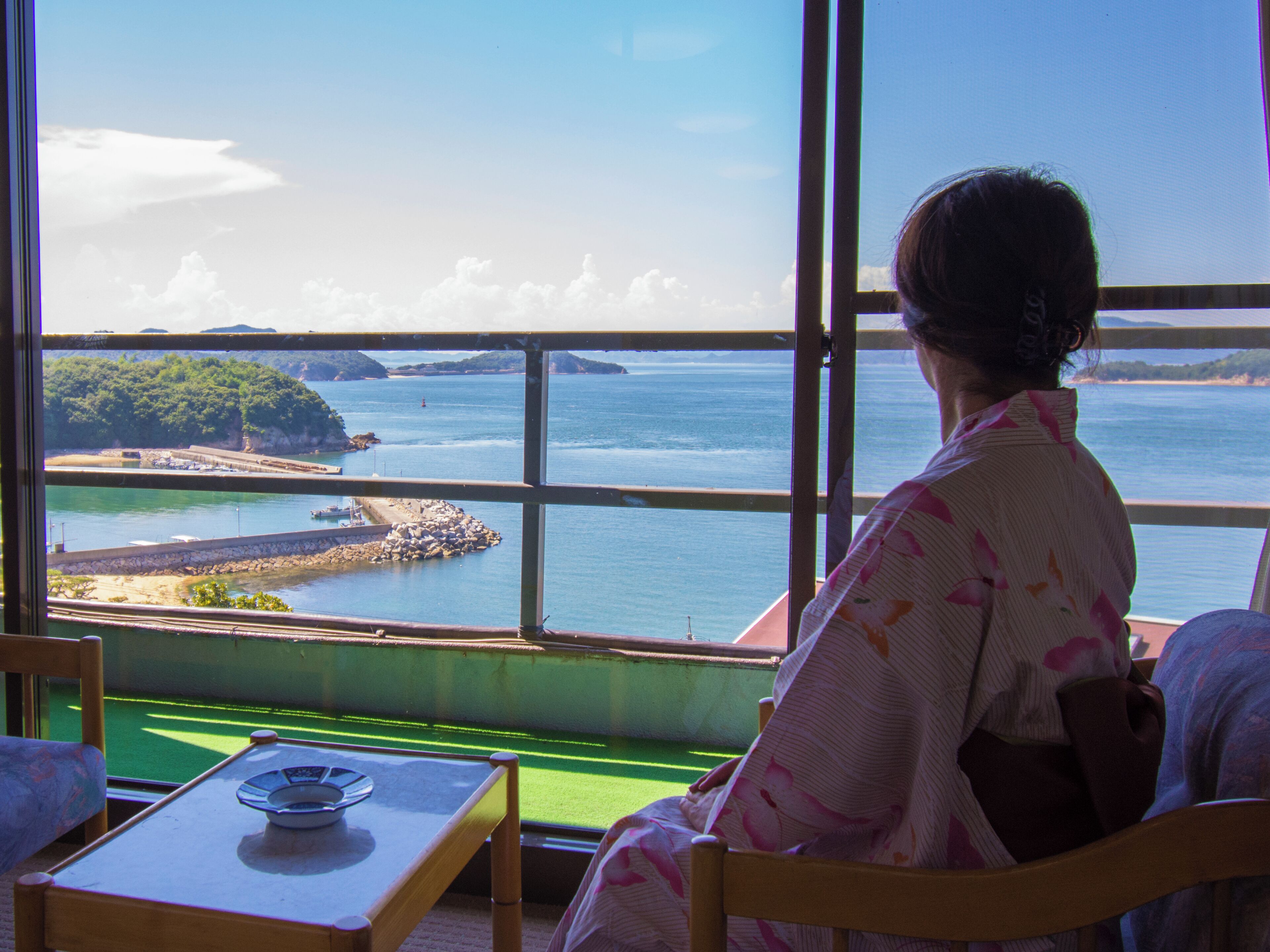A room with a large window offering a view of a coastal area, a table with a plate, and a chair.