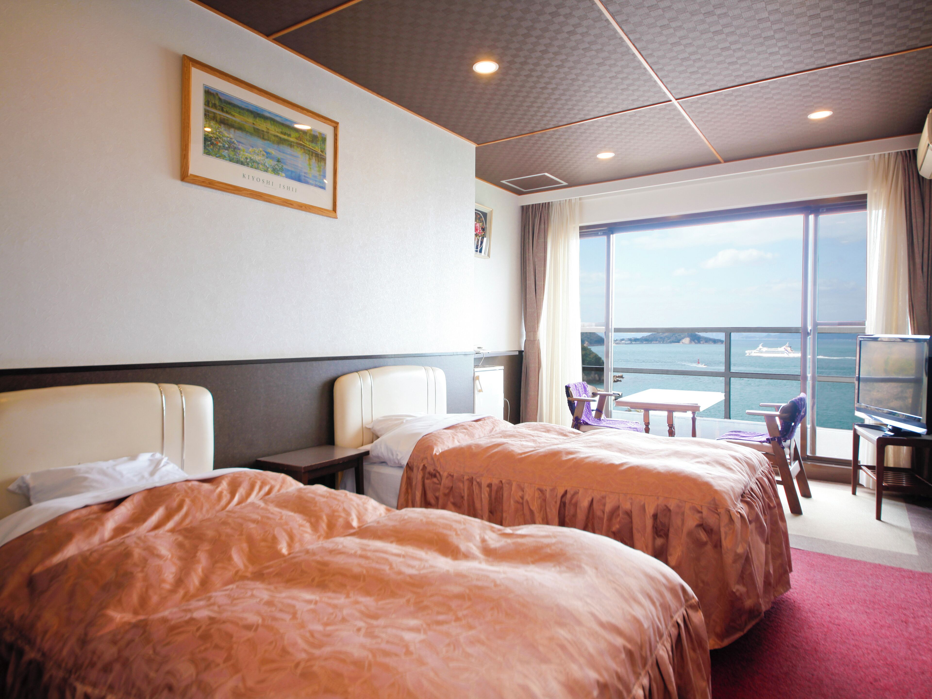 A hotel room with two beds, a desk, a chair, a television, and a large window with a view of the sea.