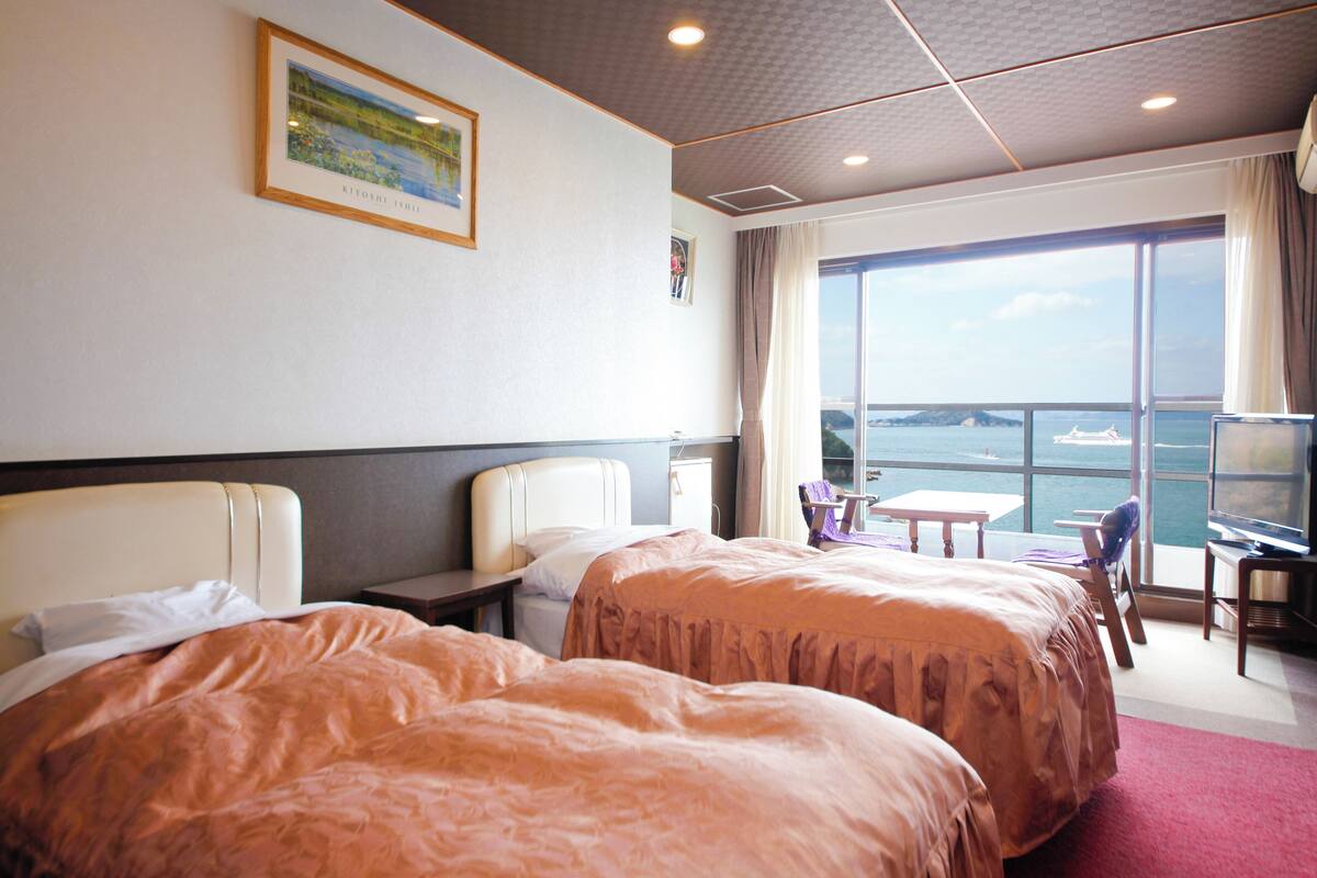 Ein Hotelzimmer mit zwei Betten, einem Schreibtisch, einem Stuhl, einem Fernseher und einem großen Fenster mit Meerblick.