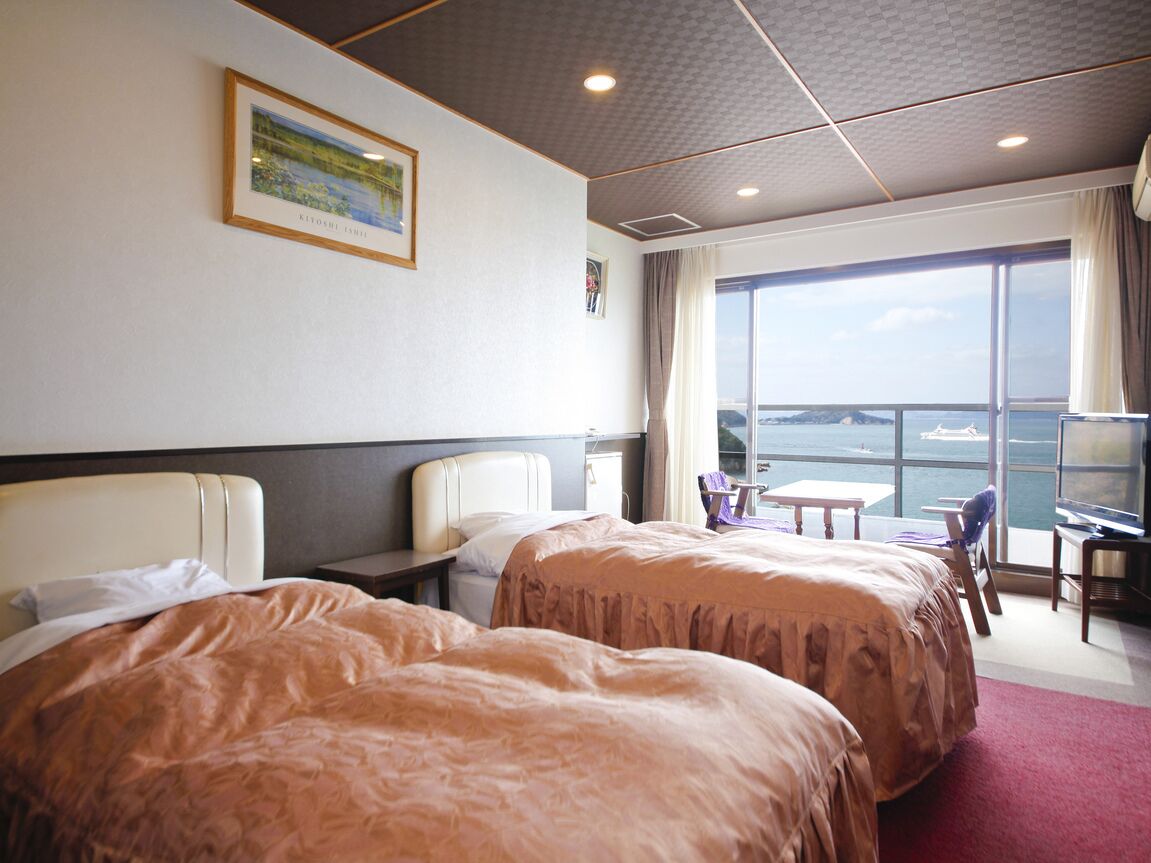 A hotel room with two beds, a desk, a chair, a window with a view of the sea, and a framed picture on the wall.