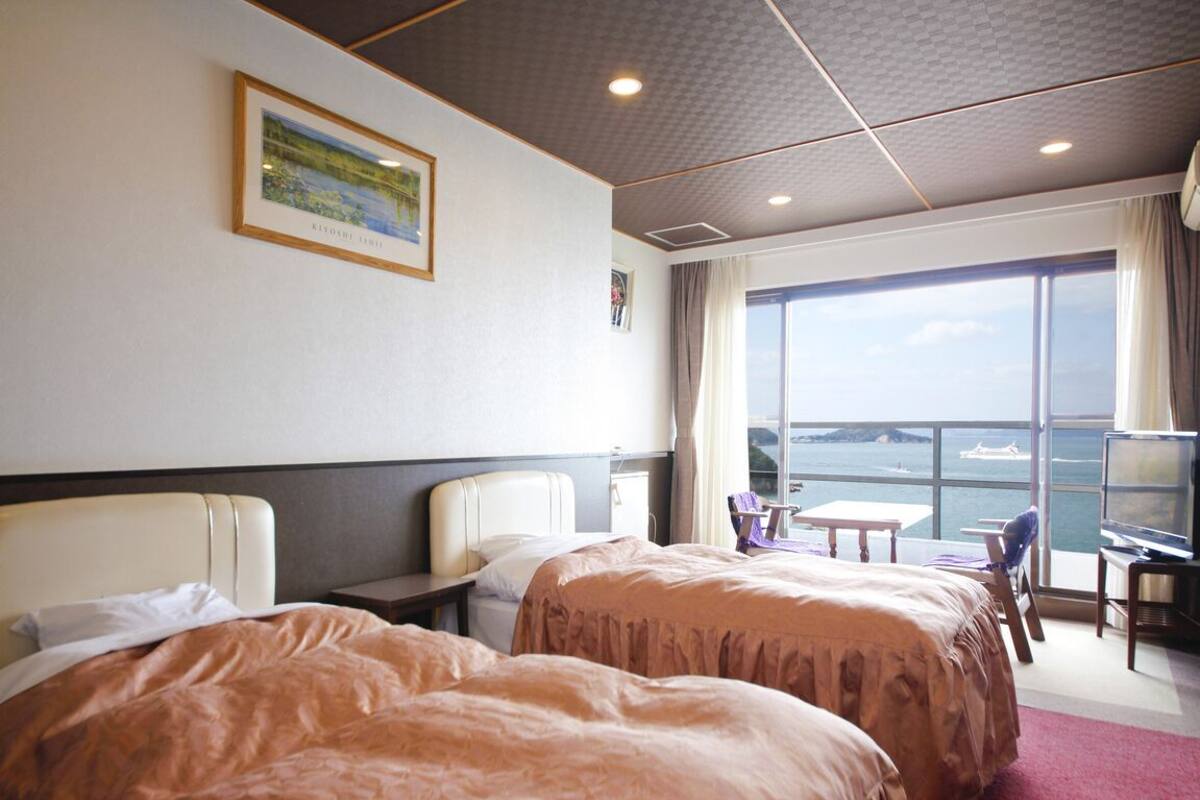 Ein Hotelzimmer mit zwei Betten, einem Schreibtisch, einem Stuhl, einem Fenster mit Meerblick und einem gerahmten Bild an der Wand.