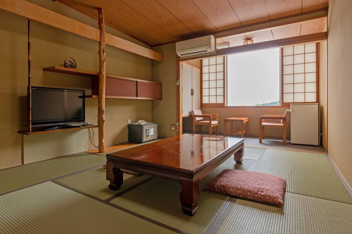 Ein traditioneller japanischer Raum mit Tatami-Bodenbelag, einem niedrigen Holztisch, einem Fernseher und einer Schiebetür zu einem Balkon.